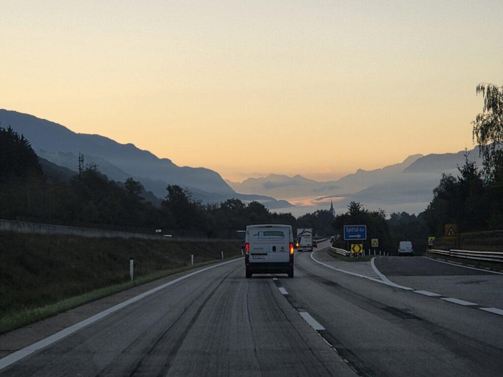 Ein Lieferwagen fährt auf einer asphaltierten Straße, im Hintergrund erheben sich Berge im Morgennebel.