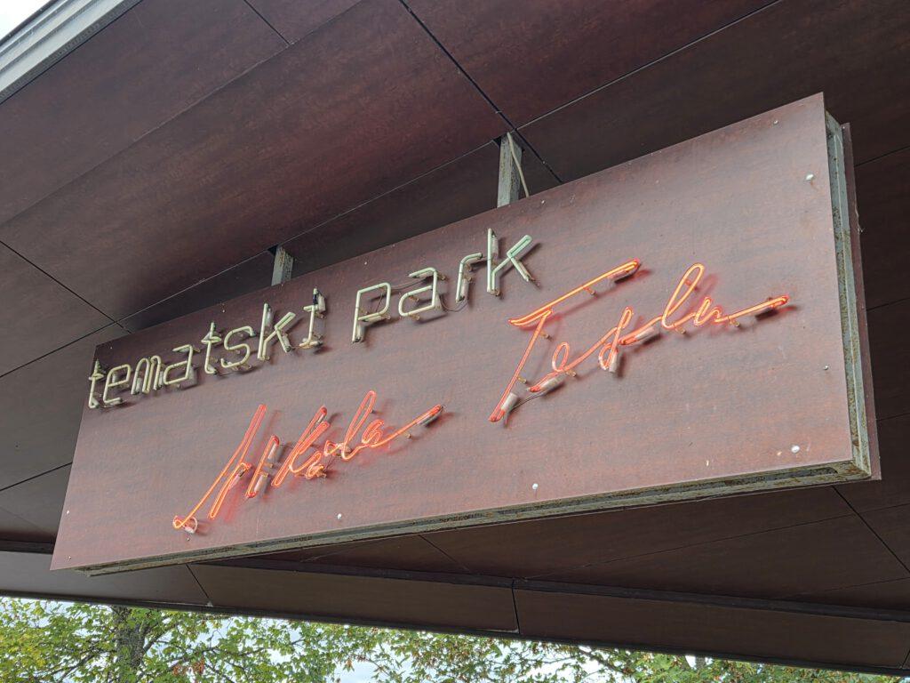 Neon-Schild mit dem Schriftzug 'tematski park' an einem grauen Holzbalken.