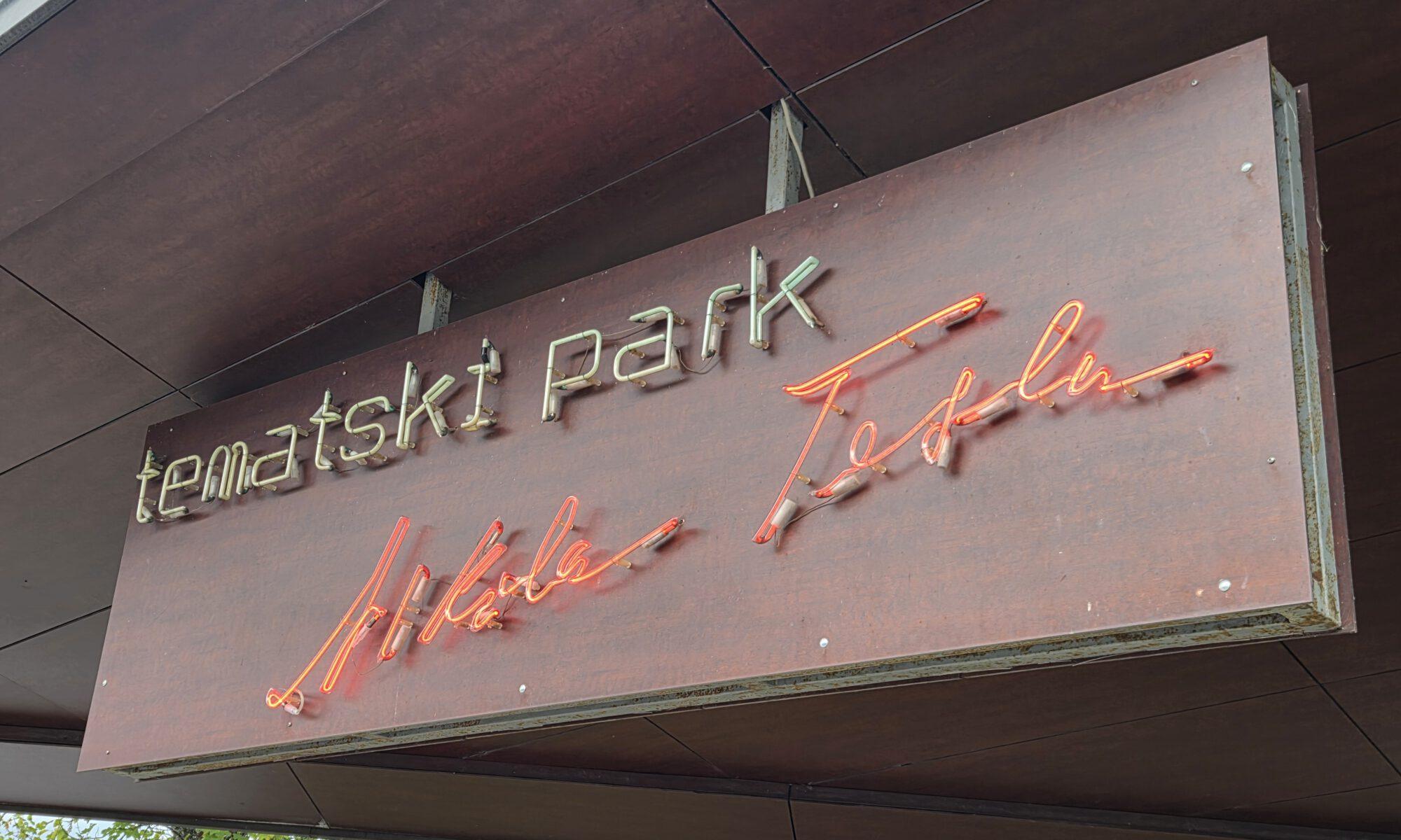 Neon-Schild mit dem Schriftzug 'tematski park' an einem grauen Holzbalken.