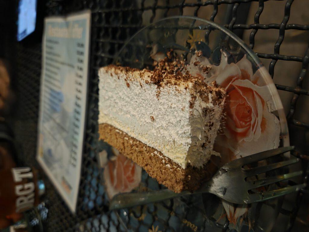 Ein Stück Käsekuchen auf einem Teller auf einem Metallgestell, daneben ein Getränk und ein Flyer.