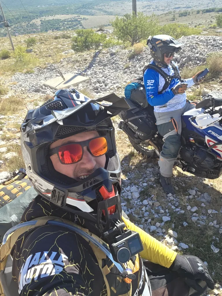 Zwei Motorradfahrer posieren lachend vor ihren Motorrädern in einer bergigen Landschaft.